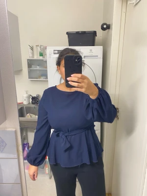 Mörkblå blus med knytskärp och dragkedja  - Snygg mörkblå blus med rund halsringning och långa puffärmar. Blusen har ett matchande knytskärp i midjan och ett lätt utställt peplumfall. Materialet ser ut att vara ett mjukt tyg som ger ett elegant intryck.