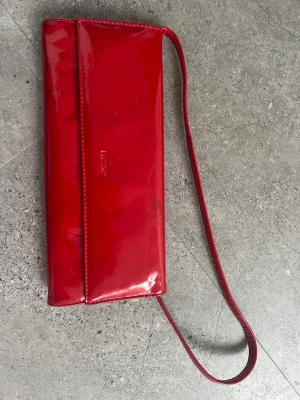 Röd lackad kuvertväska med axelrem från Picard - Snygg röd kuvertväska i glansigt lackmaterial med smal axelrem. Väskan har en enkel, rektangulär form och stängs med lock. Perfekt för att addera färg till din outfit.