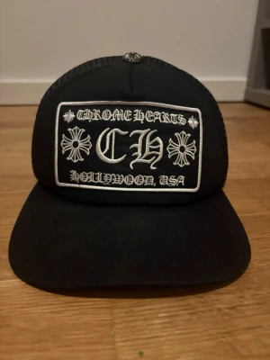 Svart Chrome Hearts truckerkeps - Säljer en svart truckerkeps från Chrome Hearts med vit broderad patch framtill där det står 'Chrome Hearts Hollywood, USA' och CH-logga. Kepsen har mesh på sidorna och justerbar snapback. Klassisk streetwear-vibe och riktigt snygg detalj till din outfit.