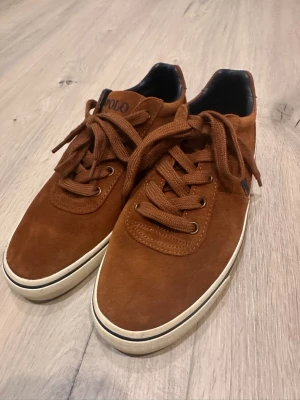 Bruna mockasneakers från Polo Ralph Lauren Storlek 41 - Snygga bruna sneakers i mocka från Polo Ralph Lauren med klassisk låg profil. Skorna har matchande bruna skosnören, vita sulor med svart rand och den ikoniska Polo-loggan broderad på sidan. Perfekta för en stilren och avslappnad look.