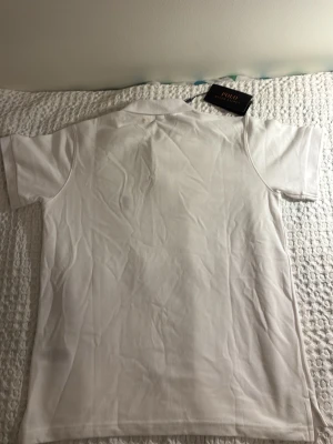 Vit pikétröja från Polo Ralph Lauren - Klassisk vit pikétröja från Polo Ralph Lauren med krage och knappar framtill. Tröjan har korta ärmar och är tillverkad i ett bekvämt bomullsmaterial. Perfekt för en stilren och avslappnad look.