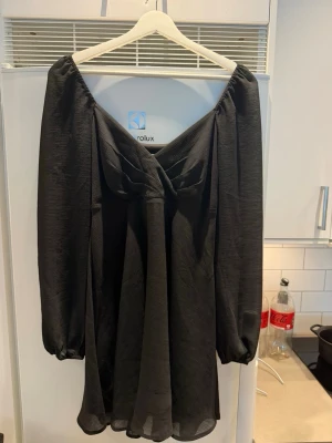 Svart omlottklänning från NA-KD - Säljer en svart omlottklänning från NA-KD x Pamela med långärmade puffärmar och v-ringad urringning. Klänningen har en luftig passform och är tillverkad i ett lätt, transparent tyg som ger en snygg drapering.