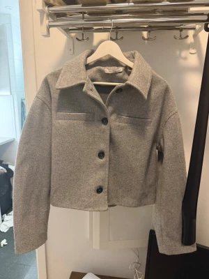 Grå overshirt från Mango - Stilren grå overshirt/kavajliknande från mango med rak passform, stora svarta knappar och klassisk krage. Overshirten har två dekorativa bröstfickor och är gjord i ett mjukt, filtliknande material som känns både trendigt och bekvämt.