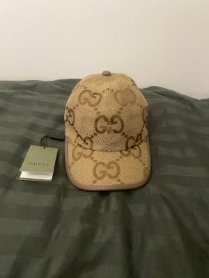 Beige keps från Gucci med GG-mönster - Snygg beige keps från Gucci med det klassiska GG-monogrammet i brunt över hela kepsen. Justerbar rem baktill i brunt läder och insida i ljus textil. Perfekt accessoar för dig som gillar exklusiv och trendig streetstyle.