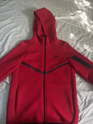 Röd Nike Tech Fleece hoodie - Nike Tech Fleece hoodie i rött med hel dragkedja, huva och svarta detaljer. Snygg svart Nike-logga på bröstet och en ficka med dragkedja på ärmen. Tillverkad i mjukt och stretchigt material som ger en clean och sportig look. Använd bara 3 gånger