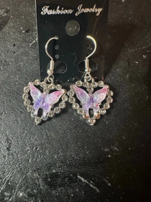 Lila fjärilsörhängen med hjärtform - Unika örhängen med lila fjärilar i mitten av ett hjärta dekorerat med glittrande stenar. Hängande modell med silvrig krok. Perfekt för dig som gillar söta och iögonfallande accessoarer. Materialet är metall och plast.