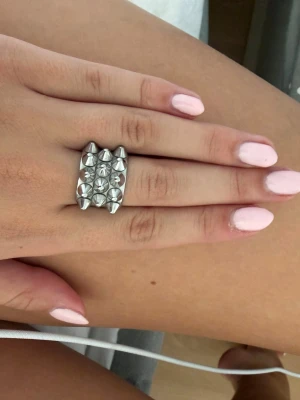 Silvrig ring med nitar och stenar - Unik silvrig ring med rektangulär form, dekorerad med små konformade nitar och glittrande stenar i ett rutmönster. Ringen har en edgy och modern look som sticker ut och passar perfekt för dig som gillar statement-smycken.