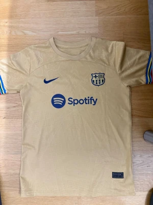 Gul FC Barcelona matchtröja Nike - Snygg gul FC Barcelona matchtröja från Nike med Spotify-logga på bröstet och klubbmärke i blått. Ärmarna har färgglada ränder och ryggen pryds av namnet Ansu Fati och nummer 10 i blått. Tillverkad i lätt och ventilerande material, perfekt för fotbollsträning.