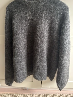 Grå fluffig stickad tröja H&M - Mysig grå stickad tröja från H&M i mohair- och ullblandning. Tröjan har en fluffig textur, rund halsringning och långa ärmar. Perfekt för kalla dagar när du vill vara både varm och snygg.