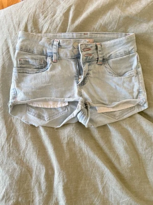 Ljusblå jeansshorts med broderi - Säljer ett par ljusblå jeansshorts med råa kanter och snygga broderade detaljer i rosa på bakfickorna. Shortsen har fem fickor, bälteshällor och dragkedja med dubbla knappar. Perfekta för varma dagar och en avslappnad stil. Kanterna har rullat upp sig lite.