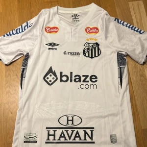 Santos FC Neymar Jr 10 matchtröja - Snygg vit Santos FC fotbollströja från Umbro med Neymar Jr och nummer 10 på ryggen. Tröjan har korta ärmar, flera tryckta sponsorer och klubbmärke på bröstet. Materialet är lätt och andas, perfekt för träning eller match. Diskreta mönster på ärmarna ger extra stil.