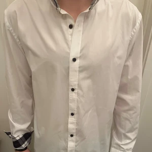 Vit slim fit skjorta Selected Homme - Snygg vit skjorta från Selected Homme i slim fit med svarta knappar och rutiga detaljer på insidan av krage och manschetter. Skjortan har lång ärm och klassisk krage, tillverkad i mjuk bomull. Perfekt för dig som gillar stilrena plagg med en twist.