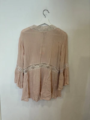 Beige blus med spets från Dotted blue - Säljer en romantisk beige blus från dotted blue med v-ringning och dekorativa spetsdetaljer vid hals, midja och ärmar. Blusen har knappar framtill och är tillverkad i ett lätt, skrynkligt bomullstyg. Ärmarna är långa och har en lätt volang vid slutet.