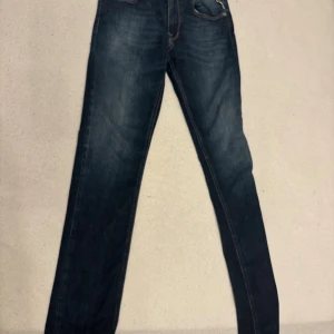 Replay mörkblå jeansbyxor 14A - Snygga mörkblå jeans från Replay med klassisk femficksdesign och kontrastsömmar. Modellen har raka ben och normal passform, med coola detaljer som slitningar vid fickan och tydliga bakfickor med dekorativa sömmar. Perfekt för dig som gillar stilrena jeans.