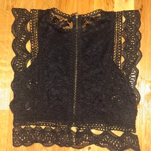 Svart spetsig ärmlös blus med dragkedja - Supersnygg svart blus i spets med dekorativa broderade kanter och ärmlös design. Toppen har en rak passform och dragkedja i ryggen som detalj. Perfekt för dig som vill ha en feminin och trendig look med extra edge.