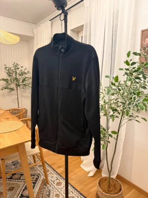 Svart ziptröja från Lyle & Scott - Svart ziptröja från Lyle & Scott med gul broderad logga på bröstet. Tröjan har hög krage, hel dragkedja och långa ärmar. Tillverkad i mjuk bomullsmix som är skön att ha på sig. Perfekt för en chill och stilren look.