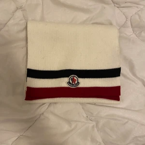 Vit stickad Moncler halsduk med ränder - Snygg halsduk från Moncler i vit stickad ull med klassiska svarta och röda ränder. Moncler-loggan är broderad framtill. Perfekt accessoar för att lyfta din vinterstil med en clean och sportig vibe.