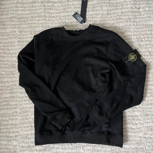 Svart Stone Island sweatshirt - Svart sweatshirt från Stone Island med klassisk rund hals och ribbade muddar. Ikonisk logotyp-patch på vänster ärm. Tröjan är i mjuk bomull och har en clean, stilren look som passar perfekt till streetwear-stilen.