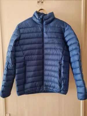 Blå dunjacka från Everest (Stadium) strl S - Blå dunjacka från Everest (Stadium) i storlek S. Jackan har horisontella quiltade sektioner, hög krage och två sidofickor med dragkedja och har innerficka. 