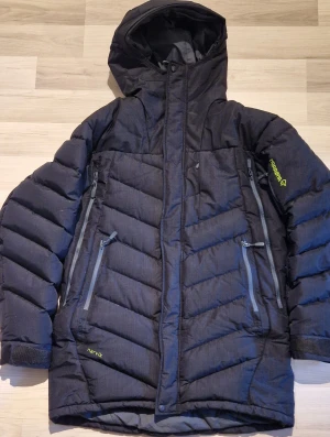 Svart dunjacka från Norrøna Narvik M - Svart dunjacka från Norrøna Narvik med huva och två stora dragkedjeförsedda fickor framtill. Jackan har quiltad design, justerbara ärmslut med kardborre och coola logodetaljer i limegrönt på ärm och rygg. Perfekt för kalla dagar och outdoor.