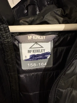 Svart pufferjacka från McKinley - Svart pufferjacka från McKinley med huva och dragkedja framtill. Jackan har horisontella quiltade sömmar, två fickor med dragkedja och en snygg, sportig look. Perfekt för kyliga dagar och enkel att matcha med din stil.