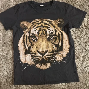 Mörkgrå t-shirt med tigertryck från Lindex - Grå t-shirt från Lindex med ett stort, detaljerat tigertryck på framsidan. Rund hals och korta ärmar. Stl 146/152 men skulle säga passar XS-S. Knappt använd 💕