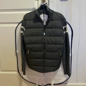 Moncler Cardigan  - Snygg grå pufferjacka från Moncler med stickade ärmar och kabelstickat mönster. Jackan har två vita ränder på ärmarna, hög krage och dubbla dragkedjor framtill. Materialmix av polyester och ull, perfekt för kyliga dagar. OBS: litet hål i högre ficka men inte som går igenom längst ner.