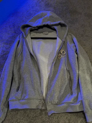 Grå hoodie Moncler - Grå hoodie från Moncler med broderade logopatchar på bröstet, dubbla dragkedjor och fickor med dragkedja. Insidan är fodrad med mjuk fleece och jackan har justerbar huva med snörning. Passar 10/10 till varenda outfit du kan tänka dig, super skön & varm och ha på sig.                        Huvud Dragkedja funkar ej, saknar dragkedja på en ficka