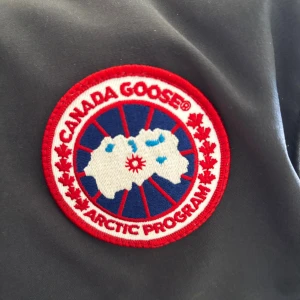 Svart dunväst från Canada Goose - Svart dunväst från Canada Goose med klassisk logga på bröstet. Västen har en quiltad design, tryckknappar framtill och två sidofickor. Materialet är slitstark polyester och dunfyllning som håller dig varm. Perfekt för lager-på-lager under kyliga dagar.