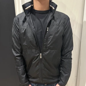 Vindjacka J.Lindberg  - Svart vindjacka från J.Lindberg. Jackan är tunn och har en inbyggd luva i kragen med dragkedja som öppnar och stänger för den. Jackan är i storlek S. Skicket är extremt bra, knappast använd. Nypris cirka 3700kr. Mitt pris 999 kr. Fråga om det finns funderingar. 