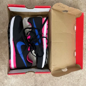 Nike sneakers svart/blå/rosa - Nike sneakers med svart mesh och skinn, blå swoosh och rosa detaljer. Snygg kontrast mellan den svarta ovandelen och den rosa/vita sulan. Rund tå, snörning och perforerad häl. Perfekt för dig som gillar färgstarka sneakers med streetkänsla.