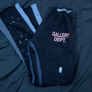 Svarta mjukisbyxor Gallery Dept. - Svarta mjukisbyxor från Gallery Dept. med rosa tryck på framsidan och coola vita färgstänk längs benen. Byxorna har elastisk midja och grå sidopaneler som ger en schysst kontrast. Perfekta för en avslappnad streetwear-look.