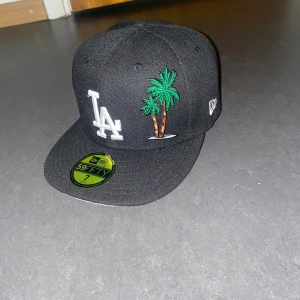 New era LA dodger keps storlek 7 - Snygg svart LA Dodgers keps från New Era 59FIFTY med broderad palm, vit LA-logga och 1988 World Series patch på sidan. Klassisk flat brim och MLB-logga bak. Perfekt för dig som gillar streetwear och baseball-vibe.