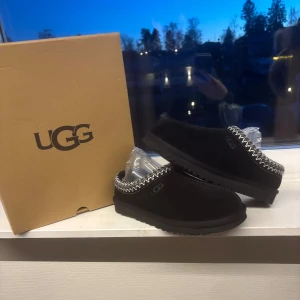 Uggs Tasman - Svarta slip-on skor från UGG med rund tå och platt sula. Skorna är tillverkade i mjuk mocka och har en snygg broderad kant i vitt och svart runt öppningen. Perfekta för dig som gillar stilren och bekväm design. Storlek 38 men passar som 37