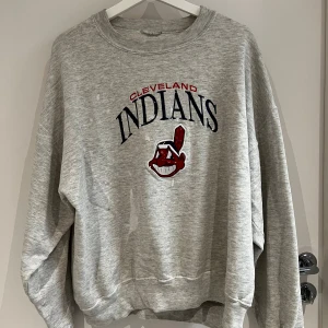 Vintage Grå Cleveland Indians sweatshirt - Säljer en grå sweatshirt med Cleveland Indians tryck i svart och rött framtill. Tröjan har rund hals, lång ärm och en loose passform. Perfekt för dig som gillar sportiga och avslappnade plagg med retro-vibe. Färgstänk sen gammalt finns sedan långt tillbaka som ger lite karaktär! 