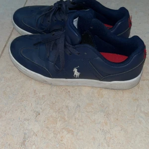 Mörkblå sneakers från Polo Ralph Lauren - Snygga mörkblå sneakers från Polo Ralph Lauren med vit logga på sidan och röd innersula. Skorna har snörning, rund tå och vit platt sula. Tillverkade i skinnliknande material med klassisk sneakerdesign och diskreta sömmar.
