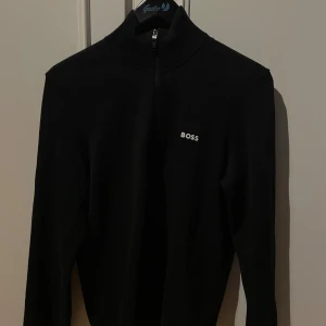 Svart half zip polotröja från BOSS - Svart polotröja från BOSS med half zip och diskret logga på bröstet. Tröjan har ribbad krage, lång ärm och mesh-detaljer vid axlarna. Snygga kontrastsömmar i grönt och vitt på insidan av kragen. Perfekt för dig som gillar stilren design.