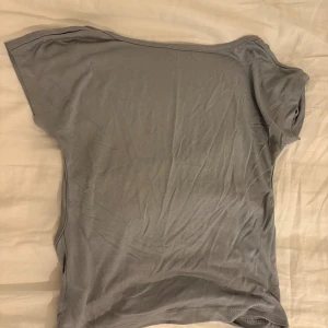 Grå omlott topp med kort ärm - Säljer en stilren grå omlott topp med korta ärmar. Toppen har en cool omlott-detalj på sidan som ger en snygg twist till den enkla designen. Materialet känns mjukt och lätt, perfekt för att matcha med jeans eller kjol. Enkel och clean look.