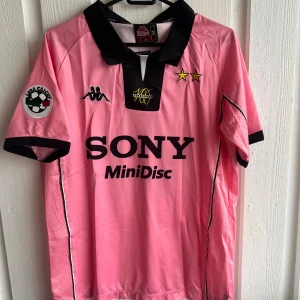 Juventus Zidane rosa fotbollströja Kappa - Juventus retro fotbollströja i rosa med svarta detaljer, tillverkad av Kappa. Tröjan har korta ärmar, svart krage och tryckta loggor från Sony och MiniDisc på framsidan. På ryggen står Zidane och nummer 21. Materialet är lätt och ventilerande polyester.