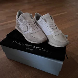 Vita Philippe Model skor - Snygga vita sneakers från Philippe Model med klassisk rund tå och platt sula. Skorna har en mix av mocka och textil, med diskret logga på sidan och vita snören. Perfekt för dig som gillar stilrena och exklusiva sneakers med fräscha detaljer. Det med kommer box och dustbag