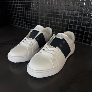 Valentino Garavani vita sneakers - Snygga vita sneakers från Valentino Garavani med bred mörkblå rand över mitten och matchande mörkblå detaljer vid snörningen. Skorna har rund tå, platt sula och är tillverkade i skinn med vita skosnören. Perfekt för dig som gillar stilrena och lyxiga sneakers.