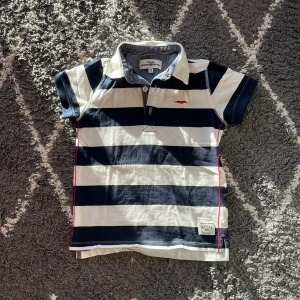 Randig pikétröja Hampton Republic - Snygg pikétröja från Hampton Republic med breda marinblå och vita ränder, röd broderad logga på bröstet och röda sömmar längs sidorna. Klassisk krage och korta ärmar, tillverkad i 100% bomull. Perfekt för en sportig och chill look.