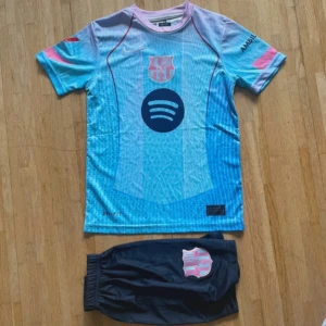 Barça Lamine Yamal fotbollströja set - Säljer ett Barcelona fotbollströja-set med Lamine Yamal #10. Tröjan är kortärmad i ljusblå och rosa toner med klubbmärke, Spotify-logga och UNHCR-tryck. Materialet är lätt och andas, perfekt för träning. Matchande mörkblå shorts med klubbmärke ingår.