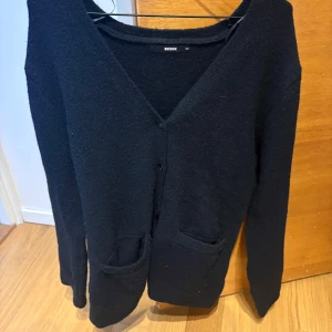 Svart kofta från Bikbok med fickor - Svart, mjuk kofta från Bikbok i storlek XS. Koftan har v-ringad hals, knappar framtill och två praktiska fickor. Långärmad och perfekt för lager-på-lager. Materialet känns stickat och ger en mysig känsla.