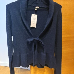 Mörkblå stickad kofta från Nelly - Supersnygg mörkblå stickad kofta från Nelly i storlek S. Koftan har långa ärmar, en bred krage och knytband framtill. Den har en lätt volangkant nedtill som ger en extra touch. Perfekt att slänga över en topp för en chill look.