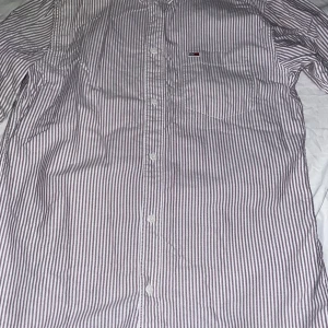 Randig skjorta från Tommy Hilfiger - Snygg skjorta från Tommy Hilfiger med smala vertikala ränder i vitt och ljuslila. Klassisk krage, bröstficka med liten logga och knäppning framtill. Perfekt för en clean och stilren look.