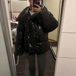 Svart oversized pufferjacka från Jannari - Säljer en svart, glansig och oversized pufferjacka från Jannari. Jackan har bred krage, långa ärmar och knappar framtill. Materialet är syntet och jackan har en cool, chunky vibe som passar perfekt när det är kallt ute. En av knapparna är lite trasiga 