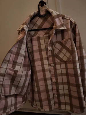 Rutig overshirt i vitt, brun och rosa - Säljer en sjukt mysig overshirt med rutigt mönster i vitt, rosa och vitt. Jackan har stora bröstfickor, knappar framtill och klassisk krage. Materialet är mjukt och fleece-liknande, perfekt för lager-på-lager. Passar dig som gillar chill och avslappnad stil.