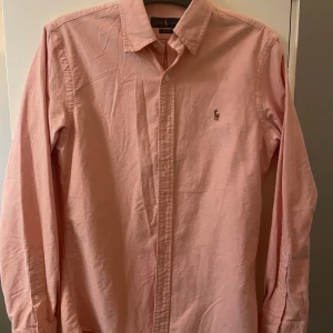 Rosa skjorta från Ralph Lauren - Säljer en snygg rosa skjorta från Ralph Lauren i slim fit-modell. Skjortan har klassisk krage, knäppning framtill och den ikoniska broderade loggan på bröstet. Tillverkad i bomull och har långa ärmar. Perfekt för dig som vill ha en stilren och fräsch look. Storlek medium med en slim fit som gör att den blir som Small