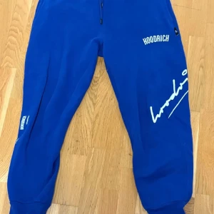 Blå Hoodrich mjukisbyxor med tryck - Snygga blå mjukisbyxor från Hoodrich med vit logotyp och tryck på benen. Byxorna har ribbade muddar vid bensluten, elastisk midja med snörning och flera fickor. Perfekta för chill eller streetwear-stil.